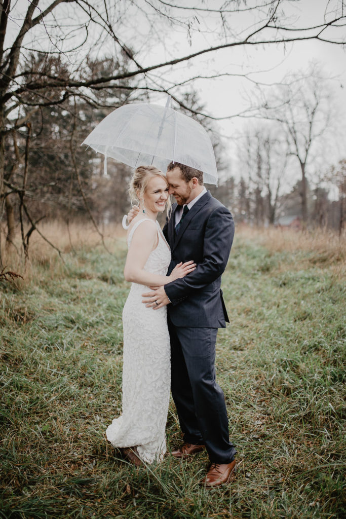 Photograph of couple standing in a field in the the rain with a clear umbrella on their wedding day by New York photographer Jessy Herman 