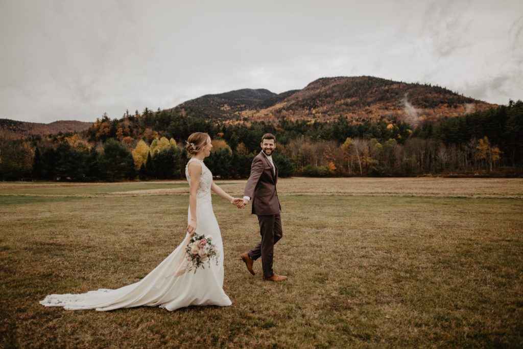 Husband and wife hold hands walking through open field with Adirondack Mountains in the background 