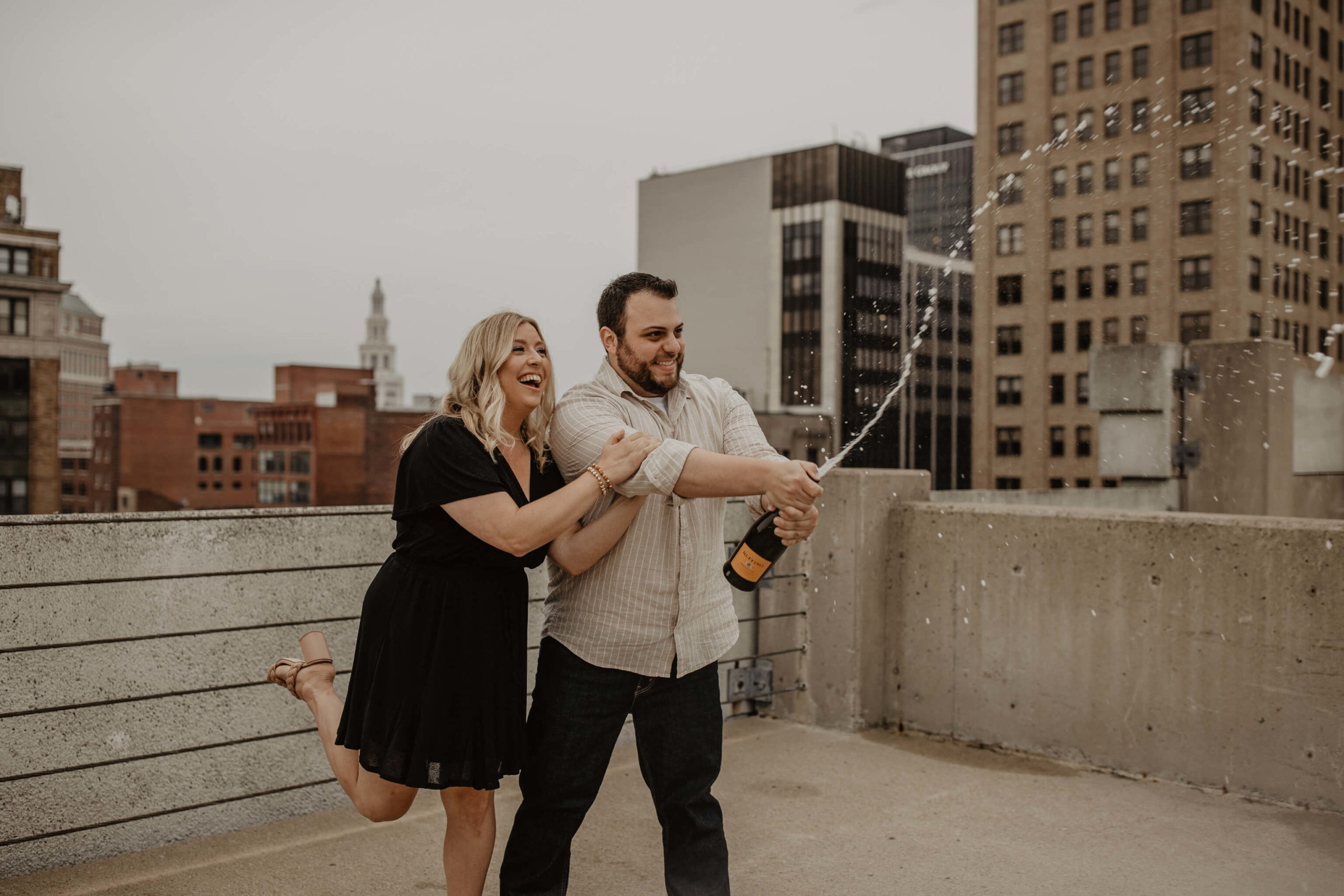 Woman in black dress and man in tan shirt pop champagne on rooftop with city skyline behind them