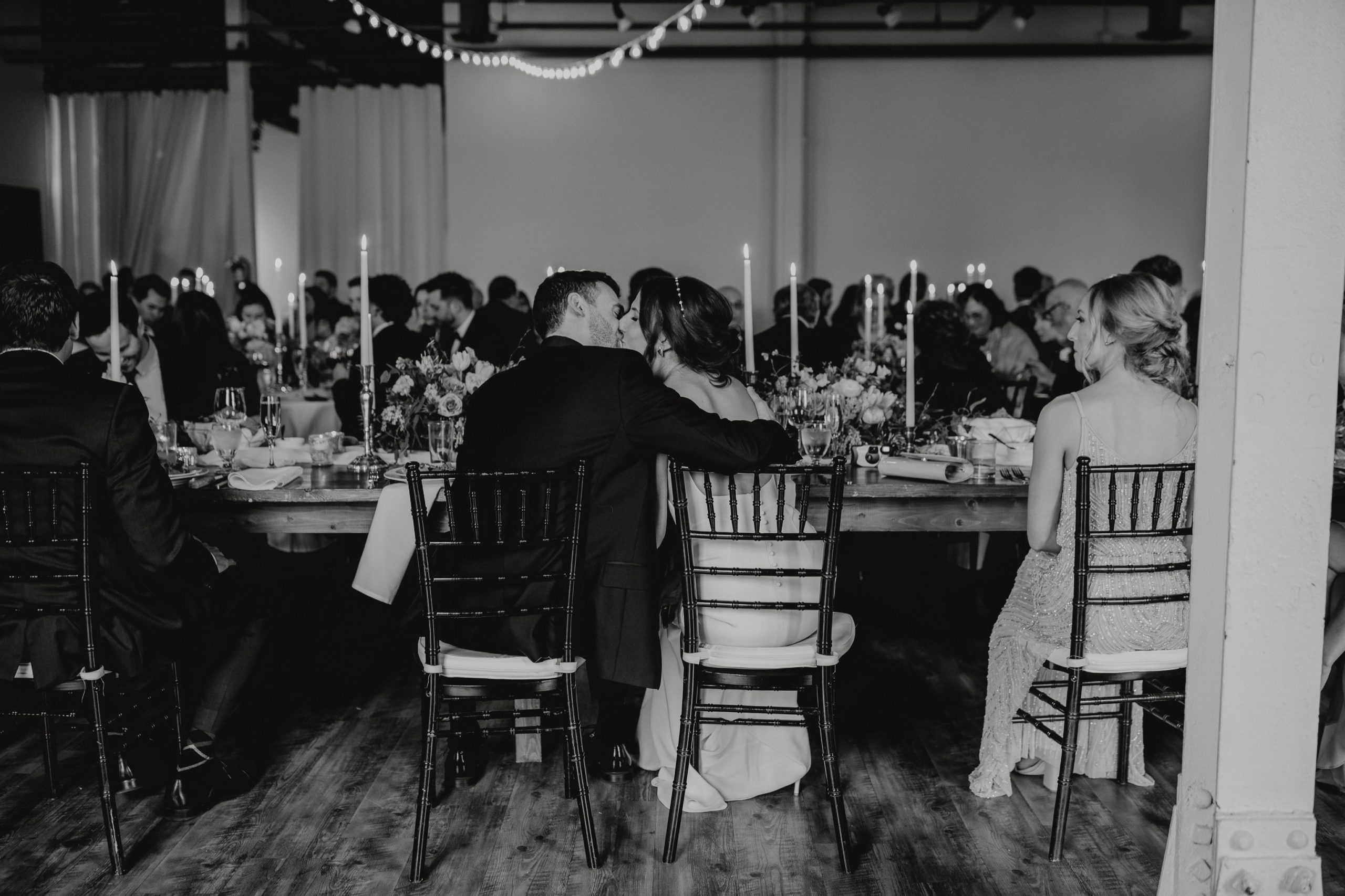 Black and white photo of bride and groom kissing from behind at their wedding reception
