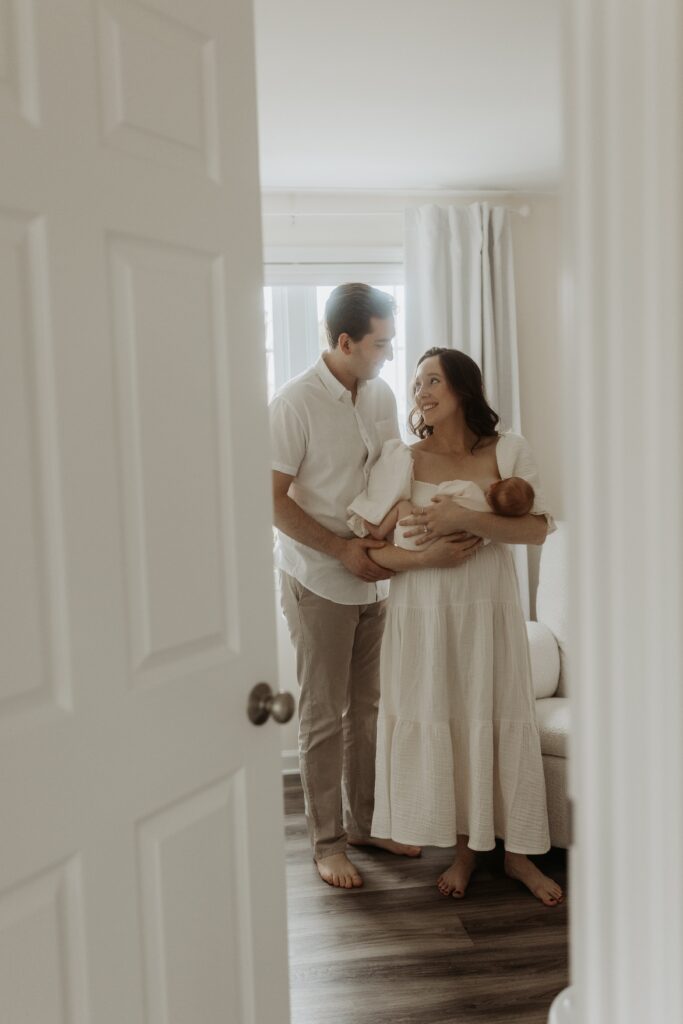 A new mom and dad smile at each other while holding their newborn baby during family photos taken by Buffalo photographer Jessy Herman Photo